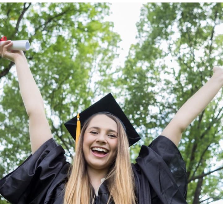 A graduate celebrating outdoors in cap and gown teacher and student wellness program